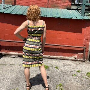 May include: A woman wearing a yellow and black striped dress with a floral pattern. The dress has thin straps and a fitted silhouette. She is standing in front of a brick wall.