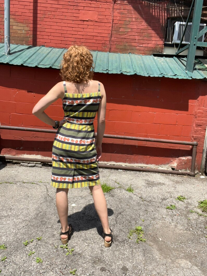 May include: A woman wearing a yellow and black striped dress with a floral pattern. The dress has thin straps and a fitted silhouette. She is standing in front of a brick wall.