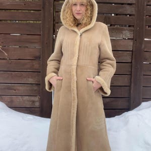 Op de afbeelding: Een lange, beige jas met capuchon, lichtbruine voering en afwerking van imitatiebont. De jas heeft een ritssluiting en zakken. De persoon staat voor een houten hek en sneeuw.