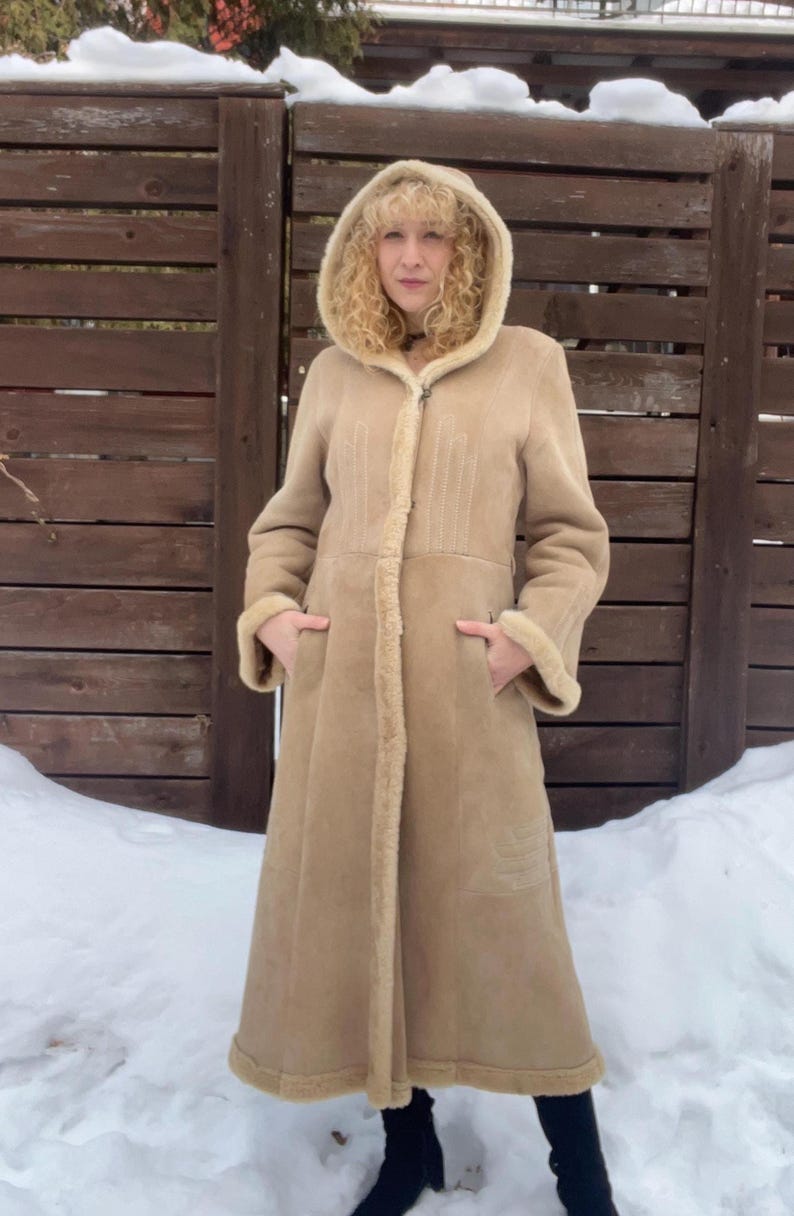 Op de afbeelding: Een lange, beige jas met capuchon, lichtbruine voering en afwerking van imitatiebont. De jas heeft een ritssluiting en zakken. De persoon staat voor een houten hek en sneeuw.