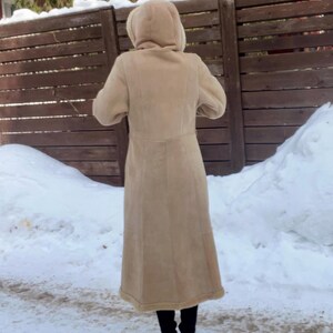 Op de afbeelding: Een lange, bruinbeige jas met een capuchon. De jas lijkt gemaakt van een zacht materiaal, mogelijk su&egrave;de of een vergelijkbare stof. De persoon draagt een zwarte broek en schoenen. De achtergrond omvat een houten hek, sneeuw en bomen.