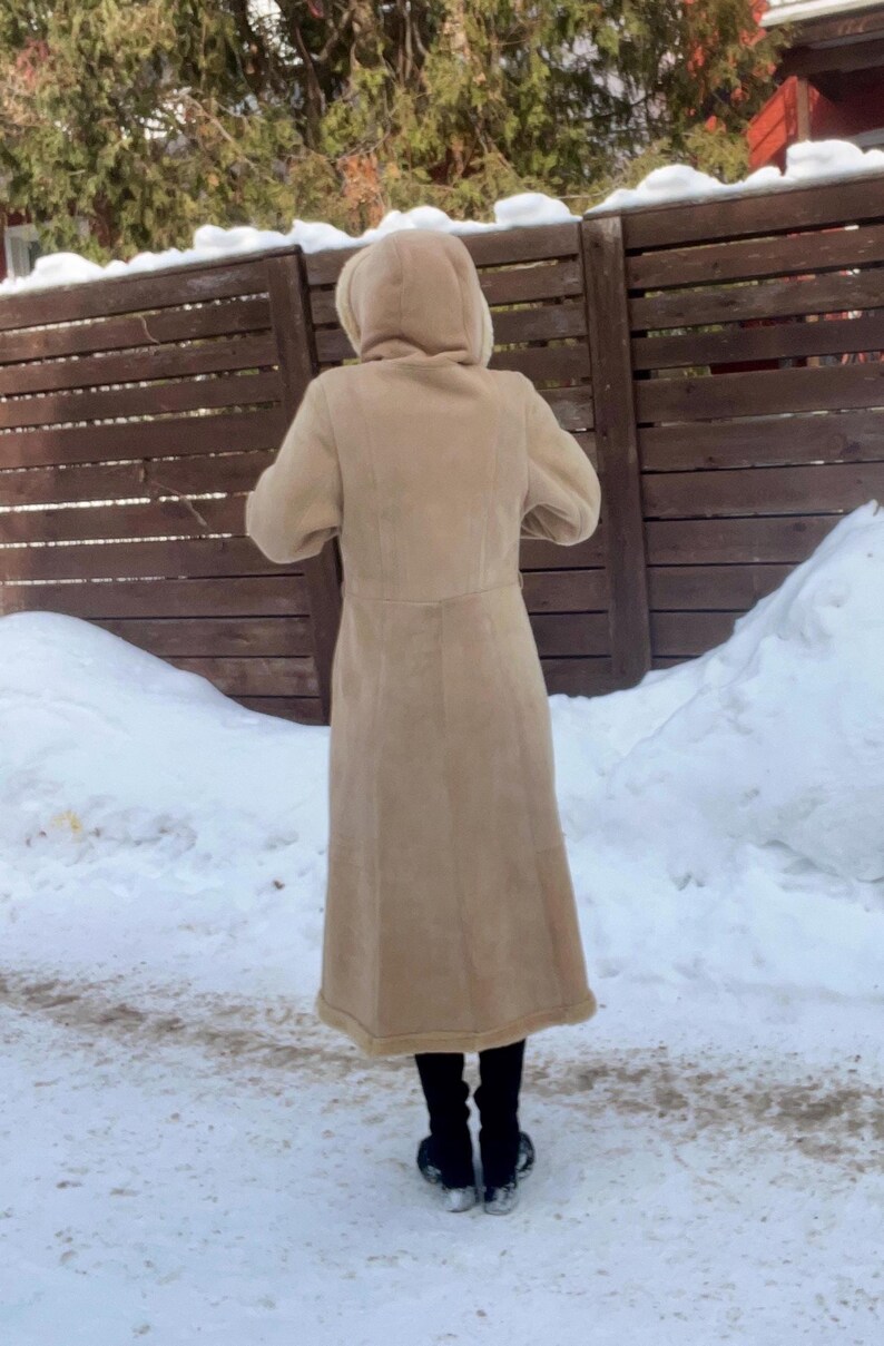 Op de afbeelding: Een lange, bruinbeige jas met een capuchon. De jas lijkt gemaakt van een zacht materiaal, mogelijk su&egrave;de of een vergelijkbare stof. De persoon draagt een zwarte broek en schoenen. De achtergrond omvat een houten hek, sneeuw en bomen.