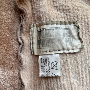 Op de afbeelding: Close-up van een beige kledingstuk met een getextureerde oppervlakte. Een rechthoekig leren label is bevestigd, met een logo en de tekst "MADE IN FINLAND". Een kleiner label met tekst in meerdere talen is ook zichtbaar.