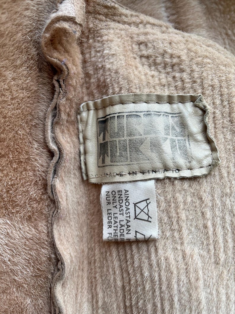 Op de afbeelding: Close-up van een beige kledingstuk met een getextureerde oppervlakte. Een rechthoekig leren label is bevestigd, met een logo en de tekst "MADE IN FINLAND". Een kleiner label met tekst in meerdere talen is ook zichtbaar.