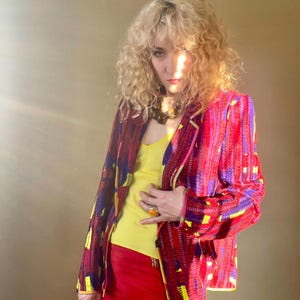 May include: A vibrant, patterned blazer in shades of red, purple, and yellow. The jacket is worn over a yellow top and paired with red pants. A large orange ring and a beaded necklace complete the look. The outfit is a bold statement.