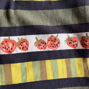 May include: A close-up of a fabric with a striped pattern in yellow, green, and brown. The fabric has a row of red strawberries with black seeds printed on it.