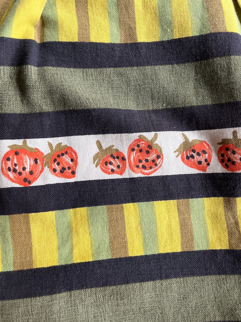 May include: A close-up of a fabric with a striped pattern in yellow, green, and brown. The fabric has a row of red strawberries with black seeds printed on it.