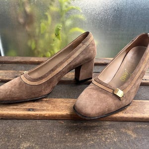 May include: A pair of brown suede pumps with a block heel. The shoes have a rounded toe and a decorative strap across the vamp. The interior of the shoe is a light tan color. The shoes are displayed on a wooden surface.
