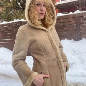 Op de afbeelding: Een beige jas met capuchon, een voering en afwerking van schapenvacht. De jas heeft een lange lengte, een knoopsluiting en zakken. Het model draagt de jas buiten in een besneeuwde omgeving.