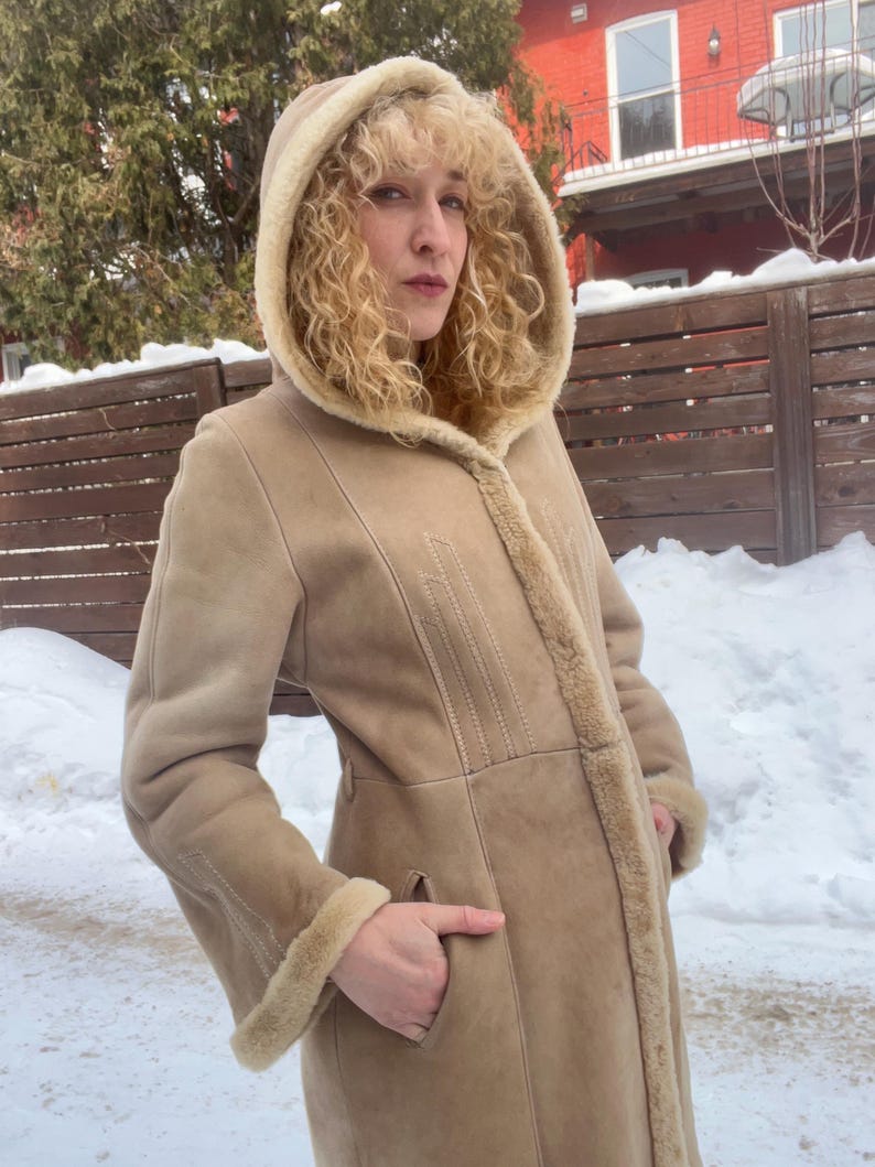 Op de afbeelding: Een beige jas met capuchon, een voering en afwerking van schapenvacht. De jas heeft een lange lengte, een knoopsluiting en zakken. Het model draagt de jas buiten in een besneeuwde omgeving.