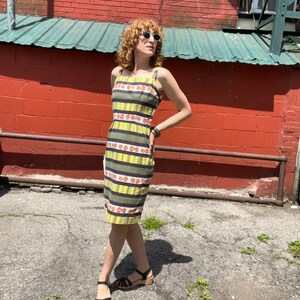 May include: A woman wearing a yellow and green striped dress with a floral pattern. The dress has thin straps and a fitted silhouette. She is wearing black sandals with a wooden heel.