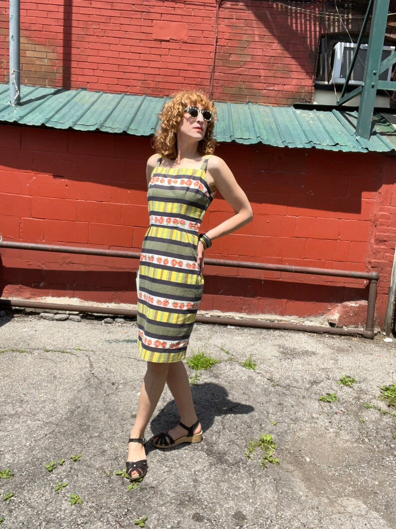 May include: A woman wearing a yellow and green striped dress with a floral pattern. The dress has thin straps and a fitted silhouette. She is wearing black sandals with a wooden heel.