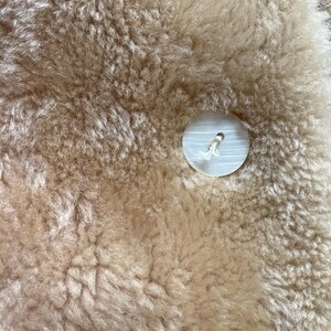 Op de afbeelding: Close-up van een beige schapenvachtkleed met een grote, ronde, witte knoop die in het materiaal is genaaid. De schapenvacht heeft een dikke, getextureerde uitstraling. De knoop heeft een klein gaatje in het midden en is bevestigd met witte draad.