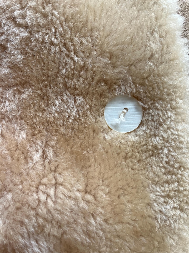 Op de afbeelding: Close-up van een beige schapenvachtkleed met een grote, ronde, witte knoop die in het materiaal is genaaid. De schapenvacht heeft een dikke, getextureerde uitstraling. De knoop heeft een klein gaatje in het midden en is bevestigd met witte draad.