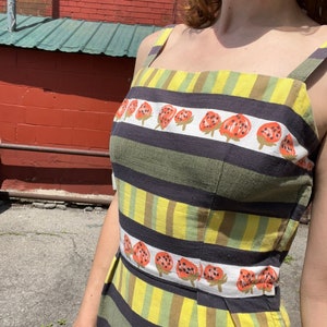 May include: A sleeveless dress with a striped pattern in black, green, and yellow. The dress has a row of red and black strawberries printed across the front.