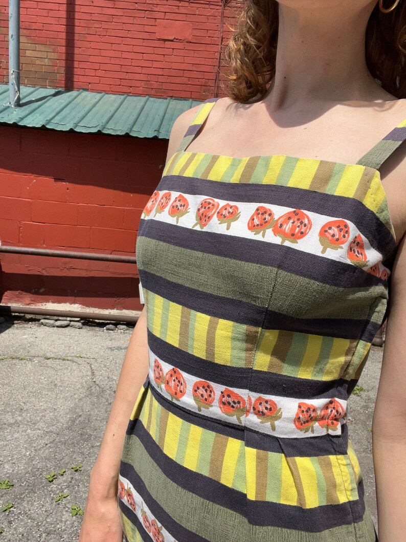 May include: A sleeveless dress with a striped pattern in black, green, and yellow. The dress has a row of red and black strawberries printed across the front.