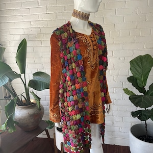 May include: A long-sleeved, burnt orange velvet tunic featuring intricate embroidery and a decorative collar, layered with a long, open-front vest constructed from a dense, multi-colored patchwork of small, circular fabric yo-yo embellishments.