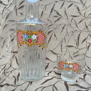 May include: A clear glass decanter with a gold rim and floral pattern, and a matching clear glass shot glass with a gold rim and floral pattern.