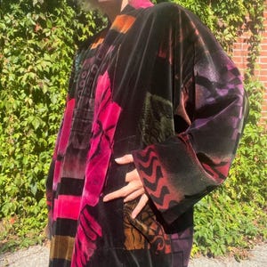 May include: A long-sleeved velvet coat featuring a dark black base with vibrant, multi-coloured abstract patterns in shades of hot pink, deep purple, and orange. The garment has a relaxed fit, wide sleeves, and a visible pocket on the side panel.