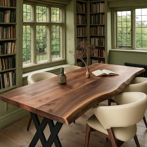 May include: A live edge walnut wood dining table with a black metal X-shaped base, accompanied by four cream-coloured upholstered chairs. On the table sits a small glass bottle, a dried plant in a vase, and an open book.