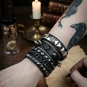 May include: A stack of five black wristbands worn on an arm. The set includes two bands with silver-toned pyramid studs, one band with silver-toned conical spikes, one band with a silver-toned metal chain, and one band with silver-toned round studs.