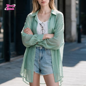 May include: A light green, long-sleeved, button-down shirt made of a sheer, textured fabric, worn over a white tank top with blue denim shorts. The shirt features a collar and a chest pocket. The logo Grace Stitch is visible in the top left corner.