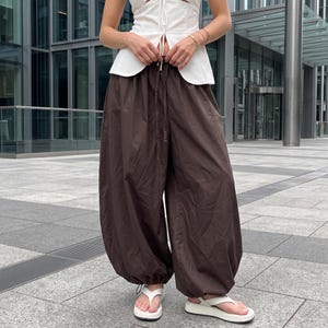 Könnte beinhalten: Dunkelbraune, weit geschnittene Hose mit hoch sitzendem Gummibund und Kordelzug. Die Hose hat eine voluminöse Silhouette mit weiten Beinen, die an den Knöcheln durch verstellbare Kordelzüge gerafft sind und so einen ballonartigen Effekt erzeugen.