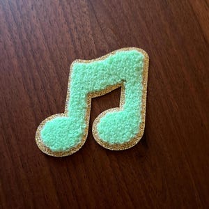 May include: A musical note patch featuring a textured, light green fuzzy center surrounded by a shimmering gold glitter border. The design is shaped like a standard eighth note with a rounded base and a stem, providing a tactile appearance.