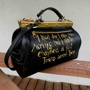 May include: Black leather doctor bag with gold hardware and a gold inscription that reads "I knew who I was this morning, but I have changed a few times since then."