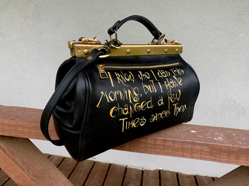 May include: Black leather doctor bag with gold hardware and a gold inscription that reads "I knew who I was this morning, but I have changed a few times since then."