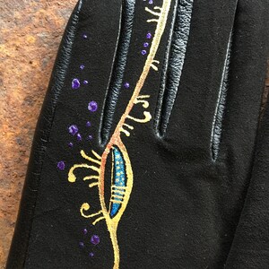 Peut inclure: Gants en cuir noir avec un motif dor&eacute; et bleu peint sur le dos de la main. Le motif repr&eacute;sente une plume stylis&eacute;e avec des points violets &eacute;parpill&eacute;s autour.