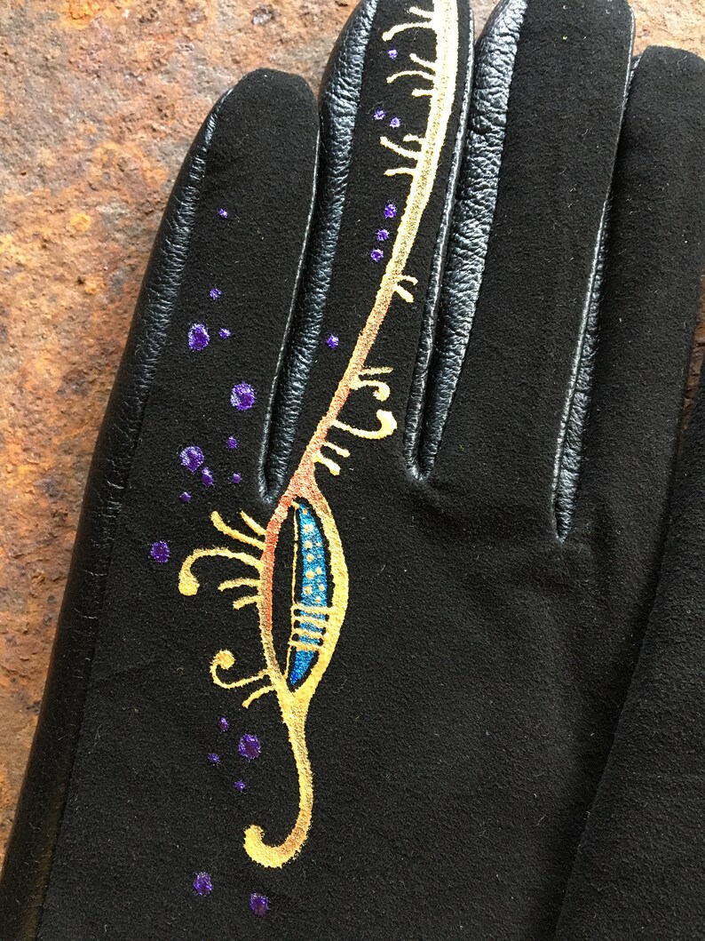 Peut inclure: Gants en cuir noir avec un motif dor&eacute; et bleu peint sur le dos de la main. Le motif repr&eacute;sente une plume stylis&eacute;e avec des points violets &eacute;parpill&eacute;s autour.
