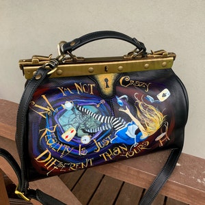May include: A black leather doctor bag with a gold metal frame and a painted design featuring Alice in Wonderland falling down a rabbit hole. The bag has a strap and the text "I'm not crazy, reality is just different than yours" is painted on the front.