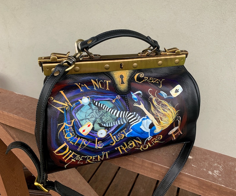 May include: A black leather doctor bag with a gold metal frame and a painted design featuring Alice in Wonderland falling down a rabbit hole. The bag has a strap and the text "I'm not crazy, reality is just different than yours" is painted on the front.