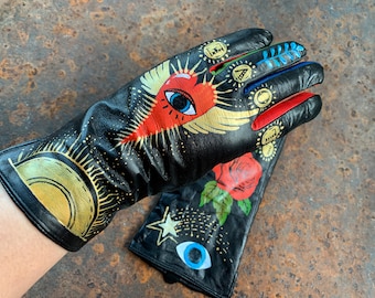 Op de afbeelding: Een paar zwarte leren handschoenen met gouden, rode, blauwe en groene geschilderde ontwerpen. De handschoenen hebben een hart met een groot oog, een halve maan, een ster, een roos en andere abstracte ontwerpen.