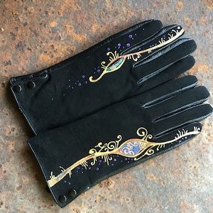 Peut inclure: Une paire de gants en cuir noir avec des motifs peints en or et violet. Les motifs sont complexes et pr&eacute;sentent une forme d'&oelig;il stylis&eacute;e.