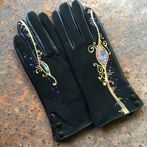 Peut inclure: Une paire de gants en cuir noir avec des motifs peints en or et violet. Les gants ont un motif d&eacute;coratif de volutes et un &oelig;il stylis&eacute;.