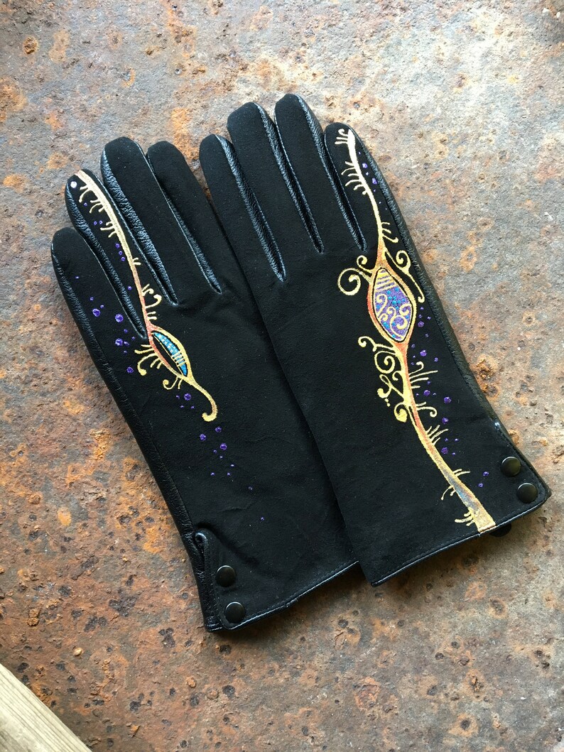 Peut inclure: Une paire de gants en cuir noir avec des motifs peints en or et violet. Les gants ont un motif d&eacute;coratif de volutes et un &oelig;il stylis&eacute;.