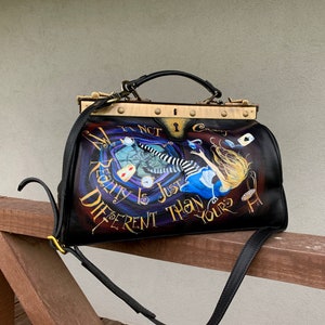 May include: A black leather doctor bag with a gold metal frame and a painted design featuring Alice in Wonderland falling down a rabbit hole. The bag has a black strap and the text "It's not crazy, reality is just different than yours".