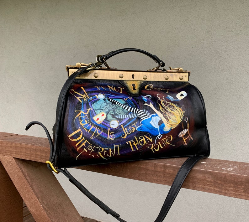May include: A black leather doctor bag with a gold metal frame and a painted design featuring Alice in Wonderland falling down a rabbit hole. The bag has a black strap and the text "It's not crazy, reality is just different than yours".