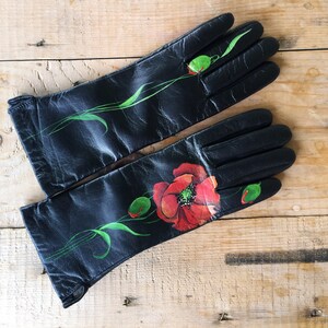 K&ouml;nnte beinhalten: Ein Paar schwarze Lederhandschuhe mit handbemaltem Mohnblumenmuster. Die Handschuhe haben gr&uuml;ne Stiele und Bl&auml;tter.