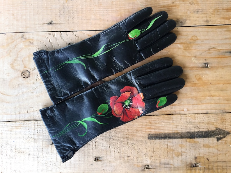 K&ouml;nnte beinhalten: Ein Paar schwarze Lederhandschuhe mit handbemaltem Mohnblumenmuster. Die Handschuhe haben gr&uuml;ne Stiele und Bl&auml;tter.