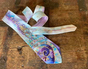Pode incluir: Uma gravata de seda com um design pintado à mão que apresenta um padrão em espiral em tons de roxo, azul e dourado. A gravata tem um fundo branco com pontos dourados dispersos.
