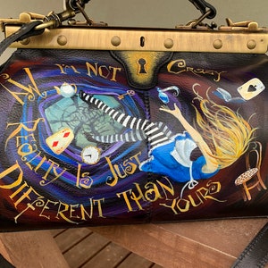 May include: A black leather doctor bag with a gold metal clasp and trim. The bag is painted with a whimsical scene of Alice falling down a rabbit hole, with the text "My Reality Is Just Different Than Yours" and "I'm Not Crazy".