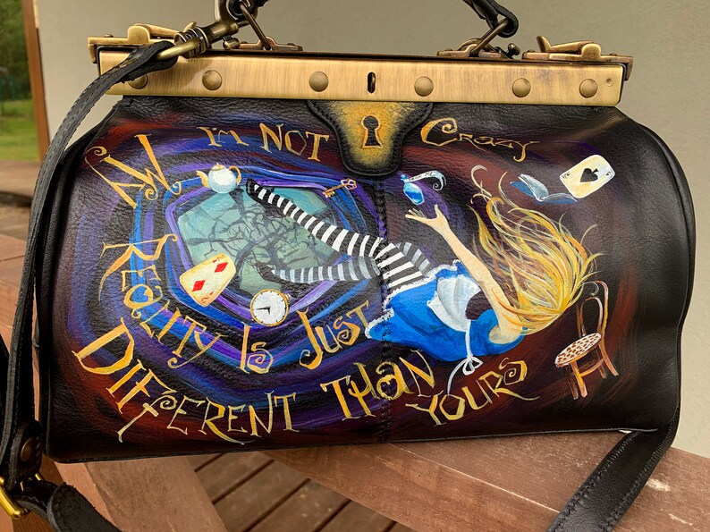May include: A black leather doctor bag with a gold metal clasp and trim. The bag is painted with a whimsical scene of Alice falling down a rabbit hole, with the text "My Reality Is Just Different Than Yours" and "I'm Not Crazy".