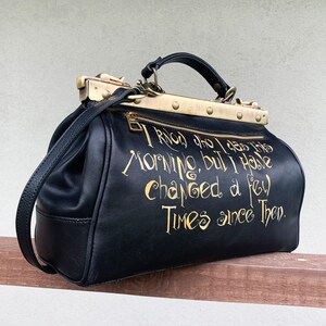 May include: Black leather doctor bag with gold hardware and a quote in gold lettering that reads "I know who I was this morning, but I have changed a few times since then."
