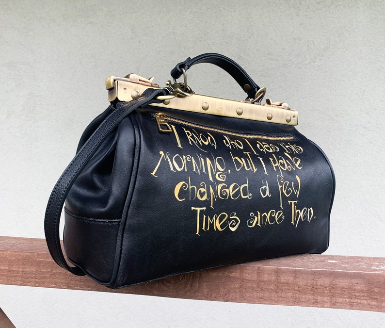 May include: Black leather doctor bag with gold hardware and a quote in gold lettering that reads "I know who I was this morning, but I have changed a few times since then."