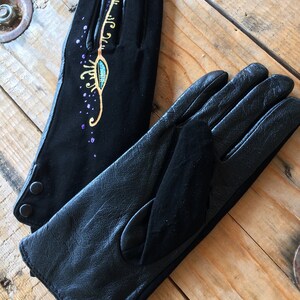 Peut inclure: Une paire de gants en cuir noir, dont l'un est orn&eacute; d'un motif dor&eacute; et turquoise. Le motif comprend un &oelig;il stylis&eacute; avec des volutes dor&eacute;es et des paillettes violettes.