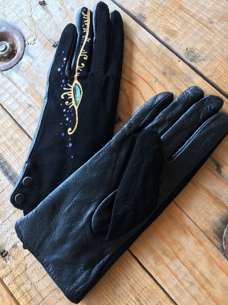 Peut inclure: Une paire de gants en cuir noir, dont l'un est orn&eacute; d'un motif dor&eacute; et turquoise. Le motif comprend un &oelig;il stylis&eacute; avec des volutes dor&eacute;es et des paillettes violettes.