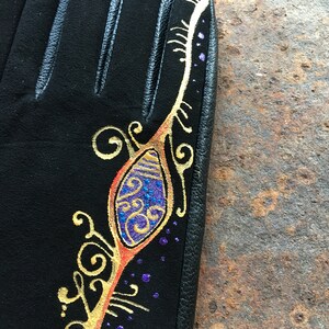 Peut inclure: Gants en cuir noir avec un motif dor&eacute; et violet sur le dos de la main. Le motif repr&eacute;sente une plume stylis&eacute;e avec des volutes et des points.